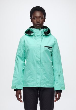 Une personne porte une veste ROXY vert menthe à capuche et à poches zippées, ainsi qu’un pantalon noir, et se tient devant un fond blanc uni.