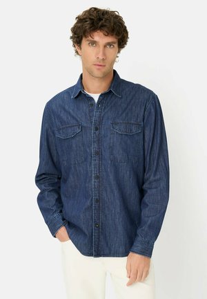 Man met krullend haar, gekleed in een donkere denimoverhemd met knopen over een wit T-shirt en witte broek, staand tegen een effen achtergrond.