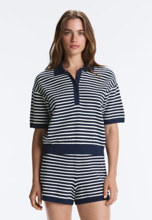 Kvinde iført navyblå og hvide vandrette stribede kortærmede strikkede polo og matchende shorts, stående mod en ensfarvet baggrund.