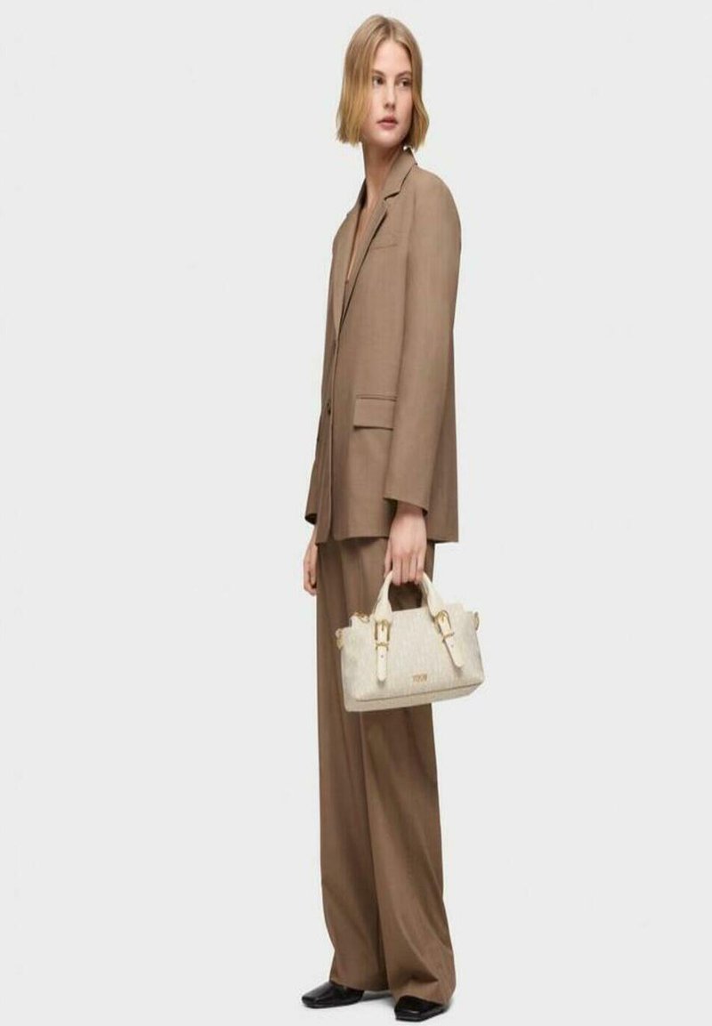 Bolso de mano beige con herrajes dorados, diseño estructurado y dos asas superiores. Combinado con un traje oversized marrón y zapatos planos negros.