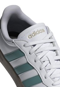 Zapatilla deportiva blanca con tres rayas en color teal, de cuero texturizado y cordones blancos. Logo dorado de "adidas" en la lengüeta. Suela de goma sutil.