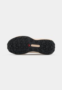 Shoe sole featuring a black rubber traction pattern with multidirectional lugs, a central cutout, and a light beige edge. Red accent included.
