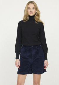 Pull en col roulé noir avec des manches ajustées, associé à des shorts en velours côtelé bleu marine avec des plis sur le devant et une fermeture à bouton.