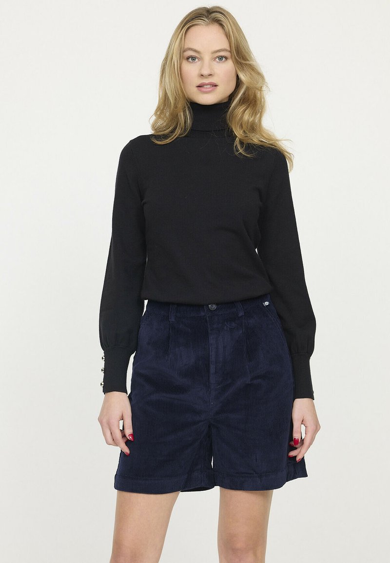 Pull en col roulé noir avec des manches ajustées, associé à des shorts en velours côtelé bleu marine avec des plis sur le devant et une fermeture à bouton.