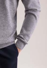 Grauer Strickpullover mit gerippten Bündchen, getragen über dunklen Marineblau-Hosen. Der Stoff wirkt weich mit einer glatten Textur und zeigt ein lässiges Design.