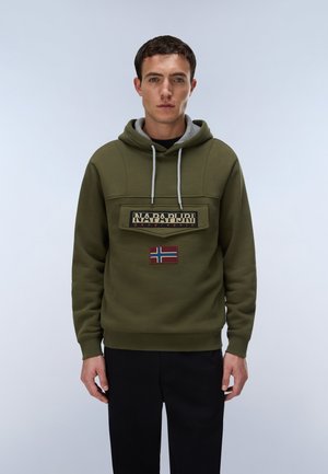 Homme portant un sweat à capuche vert olive avec poche devant, logo Napapijri et écusson du drapeau norvégien, debout devant un fond clair uni.