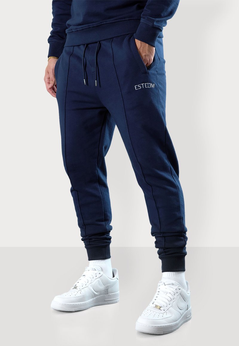 Dunkelblaue Jogginghosen mit elastischem Taillenbund, Kordelzug, seitlichen Taschen und weißer "ESTEEM" Stickerei; kombiniert mit weißen Turnschuhen.