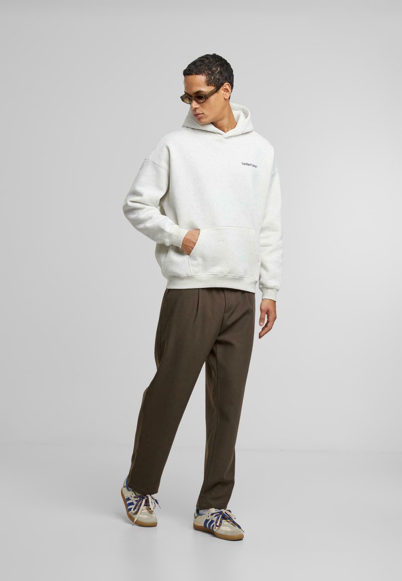 Sudadera gris con un bolsillo canguro, combinada con pantalones marrones y zapatillas multicolores, con un logo bordado en el pecho.
