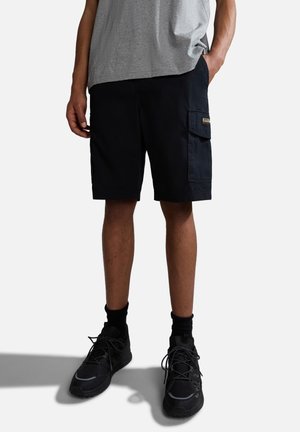 Homme portant un short cargo noir, des baskets noires et des chaussettes noires, debout avec une main dans la poche devant un fond uni.