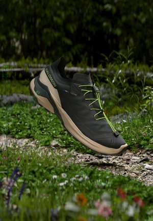 Μαύρο και μπεζ παπούτσι για τρέξιμο σε μονοπάτι Salomon με φωσφορίζοντα πράσινα κορδόνια να αιωρείται πάνω από ένα μονοπάτι με γρασίδι και βράχια σε εξωτερικό χώρο.