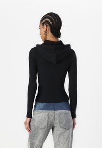 Kvinne med cornrow-fletter har på seg en tettsittende svart hettegenser og teksturerte grå bukser med blå midjebånd, vendt bort fra en hvit bakgrunn.
