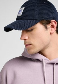 Navy corduroy cap with a textured surface and curved brim, featuring a white patch with blue logo on the front, worn with a light purple hoodie.