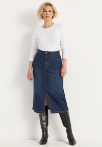 Jupe en denim bleu foncé avec fente avant, taille haute et passants de ceinture. Associée à un haut à manches longues blanc et des bottes noires jusqu'aux genoux.