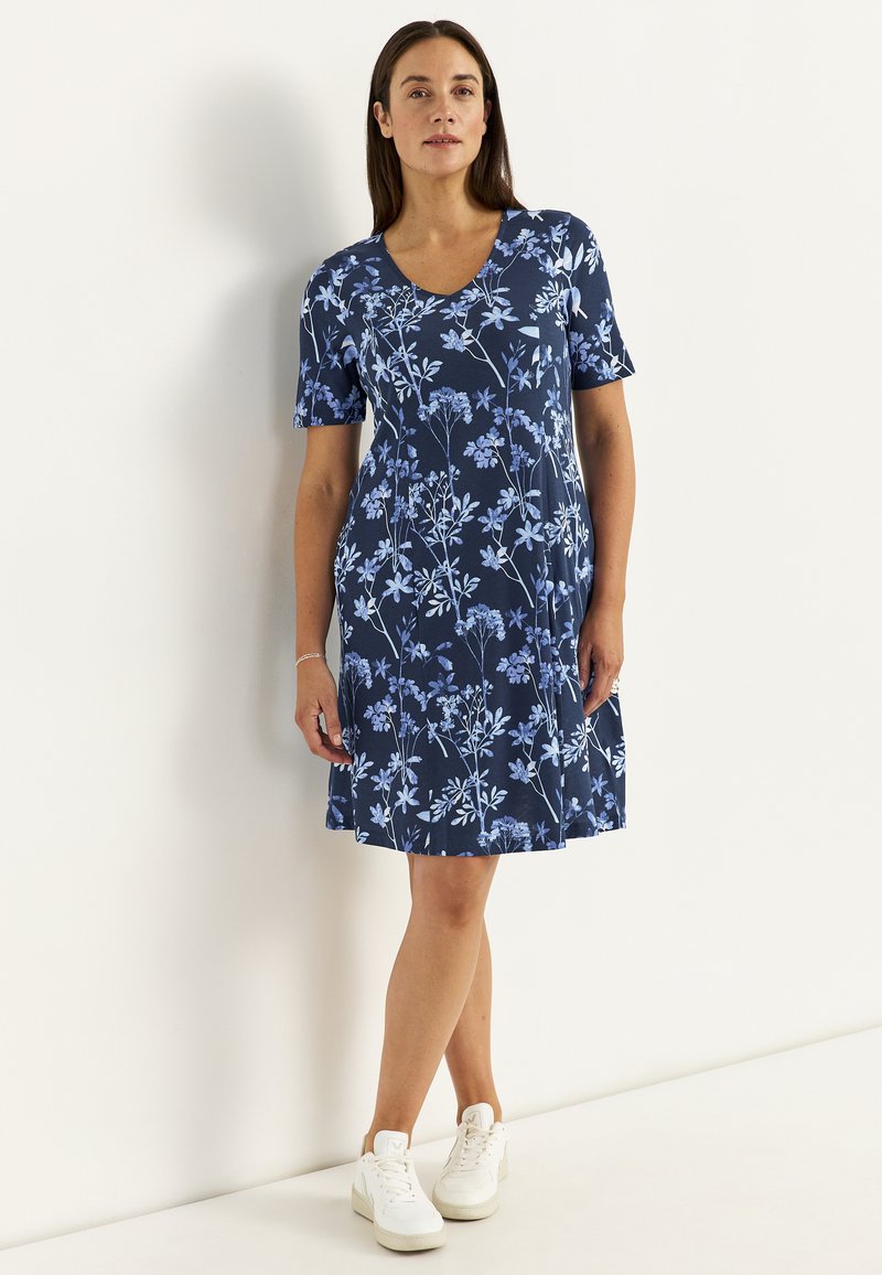 Kvinde stående iført en mørkeblå, kortærmet blomsterkjole og hvide sneakers mod en ensfarvet hvid baggrund.