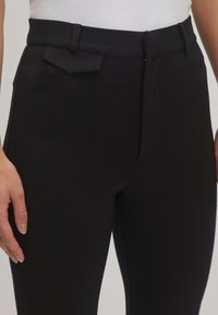 Schwarze maßgeschneiderte Hose mit glatter Textur, schmaler Passform und einem kleinen Detail an der Vordertasche. Gürtelschlaufen sind am Taillenbund sichtbar.