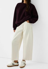 Överdimensionerad mörk lila sweatshirt, vida krämfärgade byxor med synliga sömmar, bärs med vita och svarta sneakers med gummisulor.