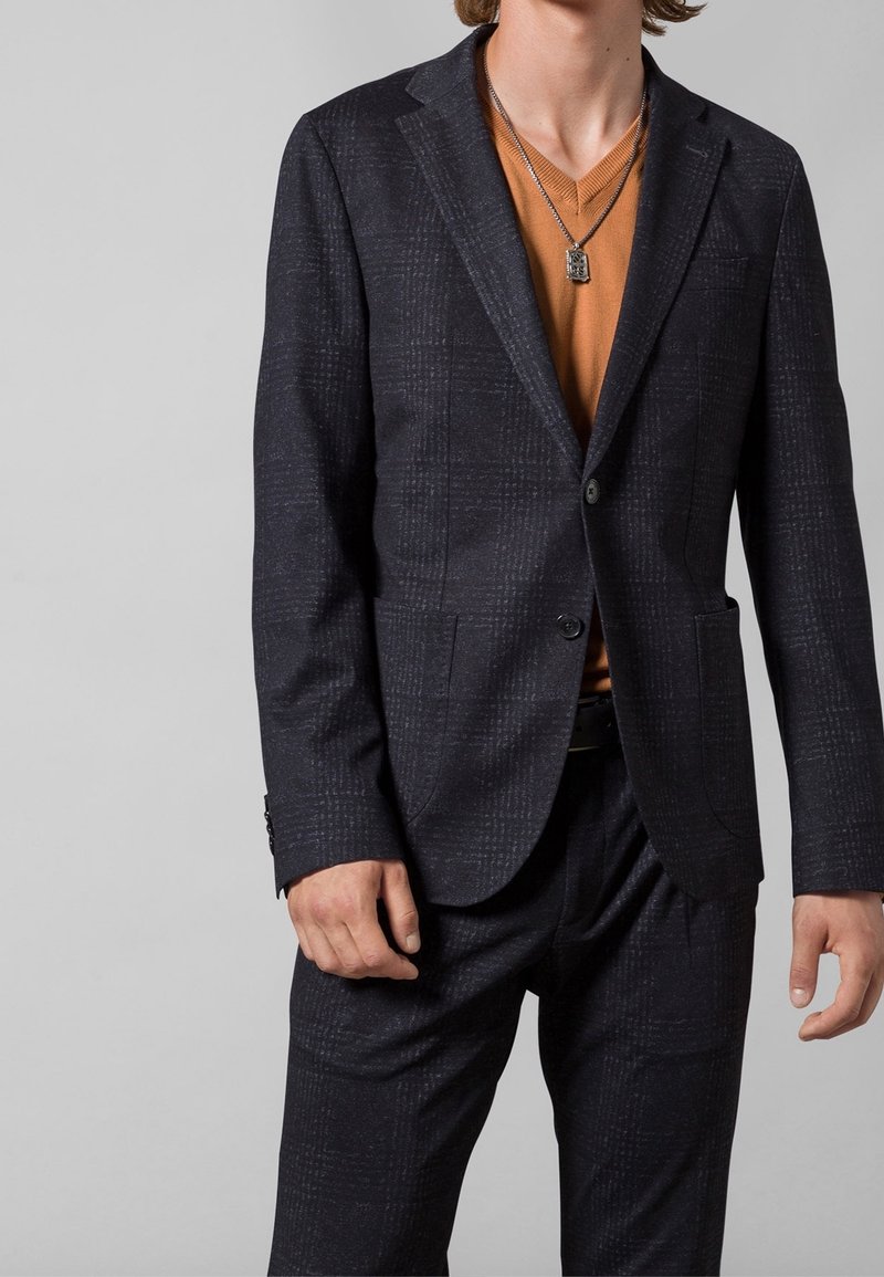 Hombre vestido con un traje de cuadros oscuros, suéter de cuello en V naranja quemado y collar de plata con colgante, de pie frente a un fondo gris liso.