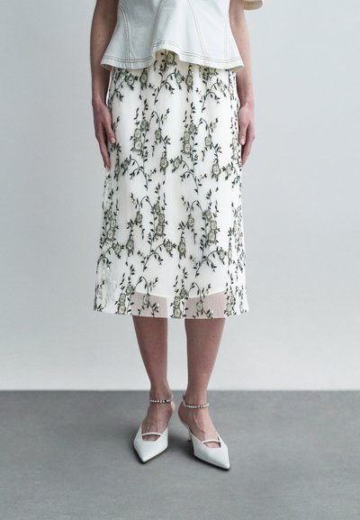 White pleated skirt with green floral embroidery, paired with white pointed-toe heels featuring ankle straps and elegant detailing.
