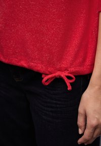 Czerwona bluzka o miękkiej fakturze z zaokrąglonym dołem, z wiązaniem w pasie. Połączona z ciemnymi dżinsami.