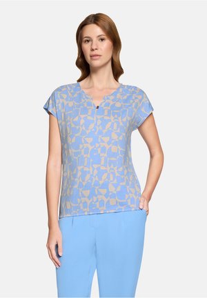 Kvinde iført en kortærmet top med geometrisk mønster i blå og beige med en lille knap ved halsudskæringen og ensfarvede lyseblå bukser.
