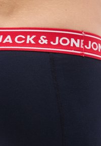 Σκούρο μπλε εσώρουχο με κόκκινη μέση που φέρει λευκά γράμματα "JACK & JON". Το ύφασμα φαίνεται λείο με ορατές λεπτομέρειες ραφής.