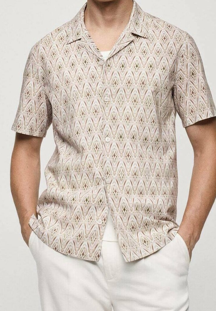 Homme portant une chemise beige à motifs à manches courtes avec les mains dans les poches d'un pantalon blanc.