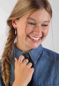 Silberne Creolen, ein strukturiertes Federdesign und gestapelte silberne Ringe. Jeanshemd mit Knopfkragen. Geflochtenes Haar.