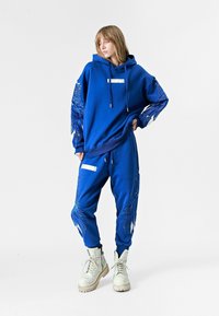 Sudadera azul y joggers a juego hechos de una mezcla de algodón, con intrincados diseños de alas azules en las mangas y los pantalones, con acentos blancos y bolsillos.