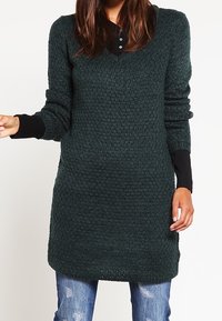 Dark green, knitted pullover dress with a textured pattern, V-neck, and long sleeves. Worn over distressed blue jeans.