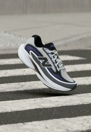 Szürke és fekete New Balance futócipő fehér talppal, légben, egy fekete-fehér gyalogátkelő felett, aszfaltburkolaton.