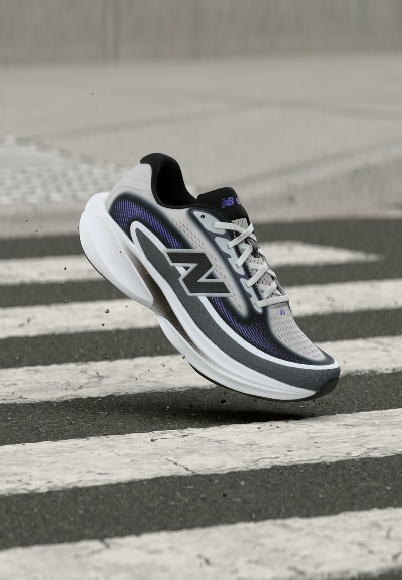 Scarpa da corsa New Balance grigia e nera con suola bianca, sospesa a mezz'aria sopra una striscia pedonale bianca e nera su pavimentazione asfaltata.