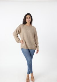Beige gebreide trui met een wijde hals en lange mouwen, gecombineerd met blauwe skinny jeans en transparante sandalen, tegen een witte achtergrond.