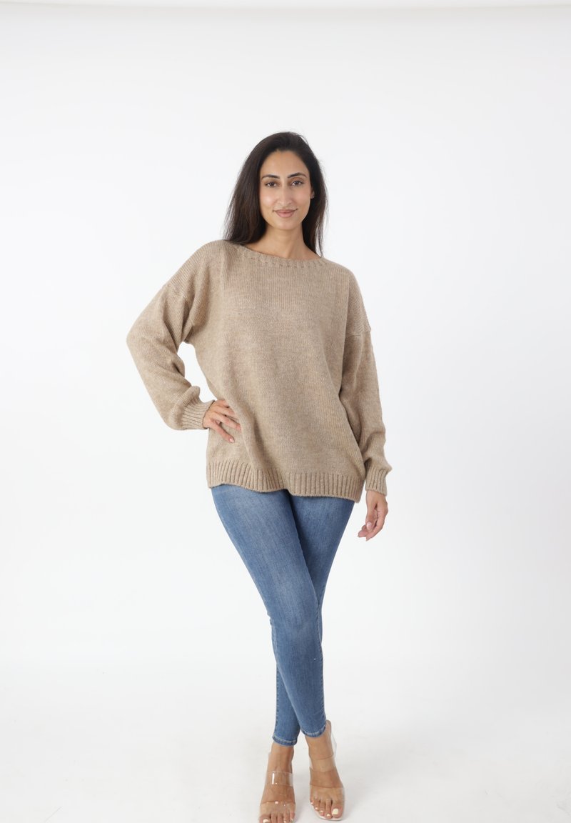 Beige gestrickter Pullover mit weitem Ausschnitt und langen Ärmeln, kombiniert mit blauen Skinny-Jeans und klaren Sandalen, vor einem weißen Hintergrund.