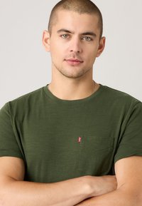 Tânăr cu părul scurt, purtând un tricou verde măsliniu cu un buzunar pe piept și o etichetă roșie mică, cu brațele încrucișate pe un fundal deschis.