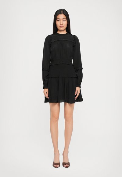 Frau, die frontal steht und ein schwarzes, langärmliges Mini-Kleid mit Rüschen sowie braune, offene High Heels trägt, vor einem weißen Hintergrund.