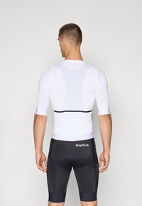 Hvit sykkeltrøye med korte ermer i strukturert stoff, med et svart elastisk bånd i livet. Bær den sammen med svarte polstrede shorts.