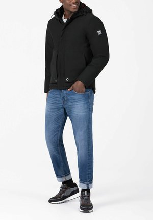 Black hooded jacket with front pockets, paired with blue rolled-up jeans. Black sneakers feature a gray accent and white sole.