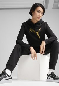 Black hoodie with gold Puma logo, paired with black joggers and black Puma sneakers featuring white accents, sitting on a white cube.