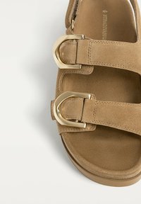 Bruine suède sandaal met twee brede banden en gouden metalen gespen, uitgerust met een gevormde voetbed en een gestructureerde zool voor comfort.