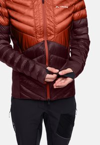 Giacca isolante rossa e bordeaux con motivo a chevron, chiusura con zip e polsini con foro per il pollice. Pantaloni neri con un ampio dettaglio della tasca laterale.