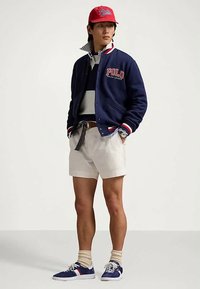 Tamsiai mėlynas universiteto stiliaus švarkas su užrašu „POLO“, balti ir raudoni akcentai, pilkas sluoksniuotas marškinėlis, smėlio spalvos šortai, mėlyni sportbačiai ir raudona kepurė.