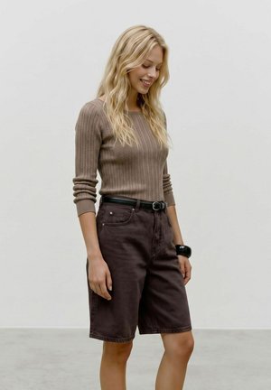 Femme aux longs cheveux blonds portant un pull en maille marron, un short marron foncé jusqu'aux genoux, une ceinture noire et un bracelet noir, souriante et regardant vers le bas.