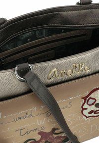 Sac en cuir brun avec un dessus en métal argenté texturé, présentant un logo doré, des motifs floraux et un texte manuscrit. L'intérieur comprend des poches.