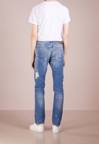 Blå denimjeans med lätt slitage, rak benstil och fem fickor. Har en logopatch på bakfickan. Bärs med en vit T-shirt.