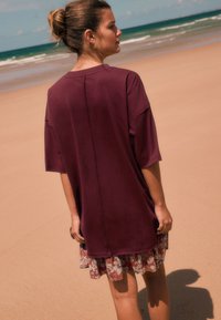 Top oversized color granate con un diseño estilizado, que presenta una costura trasera y un dobladillo floral estampado, usado en una playa de arena.