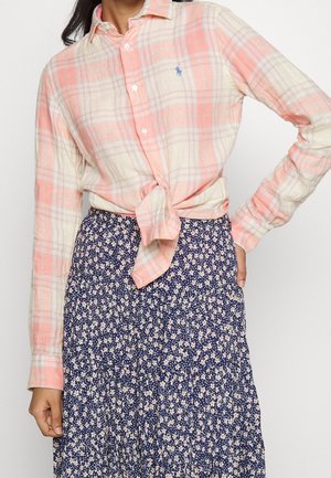Femme portant une chemise à manches longues à carreaux rose et crème nouée, avec une jupe fleurie bleu marine, debout, une main sur la hanche.