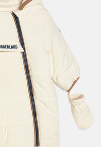 Veste doudoune de couleur crème avec un accent beige, fermeture éclair marine et logo sur la poitrine. Comprend des poignets élastiques et des moufles attachées.