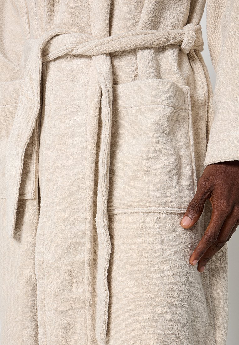 Zbliżenie dłoni odpoczywającej w pobliżu kieszeni beżowego szlafroka z frotte, zawiązanego materiałowym paskiem.