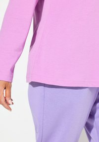 Top rosa a maniche lunghe con orlo arrotondato, abbinato a pantaloni viola. Tessuto liscio e vestibilità comoda, con dettagli di cucitura visibili.