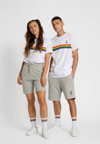 Avslappnade vita T-shirts med regnbågsspår över bröstet, i kombination med grå shorts. Båda bär regnbågssockor och vita skor.